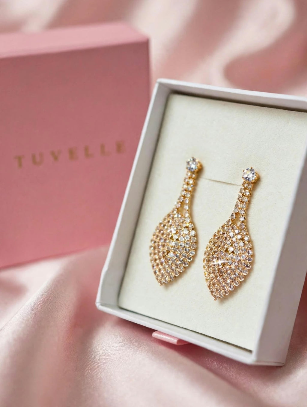 Gold-tone leaf drop earrings beside a pink TUVELLE box on pink satin