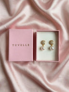 Angled view of gold-tone pearl-look drop earrings with pink TUVELLE packaging