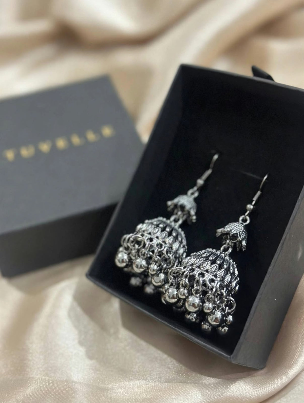 Top view of silver-tone jhumka earrings in black TUVELLE packaging on cream satin