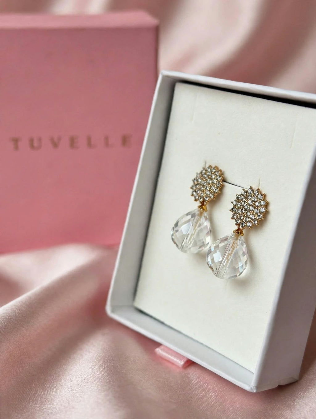 Clear teardrop earrings displayed beside a pink TUVELLE box on pink satin